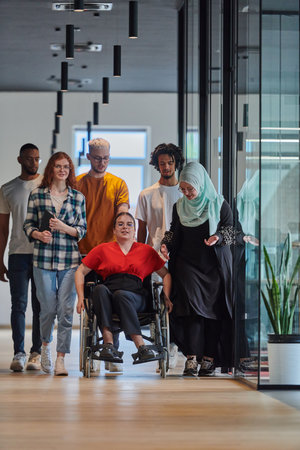 A diverse group of young business people walking a corridor in the glass-enclosed office of a modern startup, including a person in a wheelchair and a woman wearing a hijab, showing a dynamic mix of innovation and unity.の写真素材