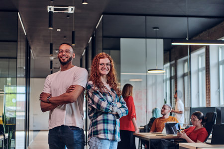 A young African American businessman and a modern businesswoman with orange hair stand side by side, arms crossed, exuding confidence and unity in a contemporary office setting, epitomizing dynamic collaborationの写真素材