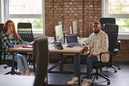 Group of colleagues, a woman with vibrant orange hair and a young African American businessman, sitting in a modern office space, symbolizing diverse collaboration and a dynamic work environment.の写真素材