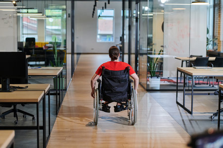 A modern young businesswoman in a wheelchair is surrounded by an inclusive workspace with glass-walled offices, embodying determination and innovation in the business worldの写真素材