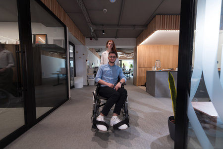 Young business colleagues, collaborative business colleagues, including a person in a wheelchair, walk past a modern glass office corridor, illustrating diversity, teamwork and empowerment in the workplace.の写真素材