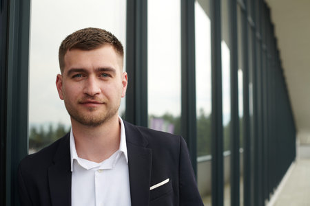 A CEO dressed in a sleek black suit stands confidently at the entrance of a modern corporate building, awaiting the start of the workday in the bustling urban environment.の写真素材
