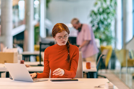 In a modern startup office, a professional businesswoman with orange hair sitting at her laptop, epitomizing innovation and productivity in her contemporary workspace.の写真素材
