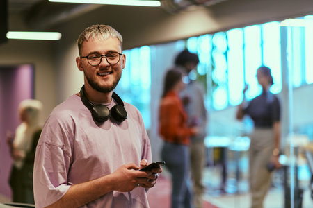 In a contemporary startup office, a modern blond entrepreneur stands, engrossed in his smartphone, epitomizing the dynamic and tech-savvy essence of the modern business world.の写真素材