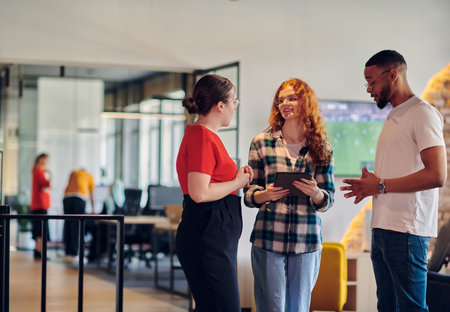 A group of young business individuals, including a girl with orange hair and an African American man, stands in a modern corporate hallway, collectively examining business progress on a smartphone, exemplifying dynamic collaboration and digital engagement.の写真素材