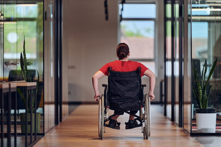 A modern young businesswoman in a wheelchair is surrounded by an inclusive workspace with glass-walled offices, embodying determination and innovation in the business worldの写真素材