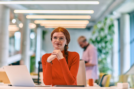 In a modern startup office, a professional businesswoman with orange hair sitting at her laptop,...