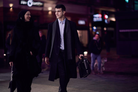 Happy multicultural business couple walking together outdoors in an urban city street at night near a jewelry shopping store window.の写真素材