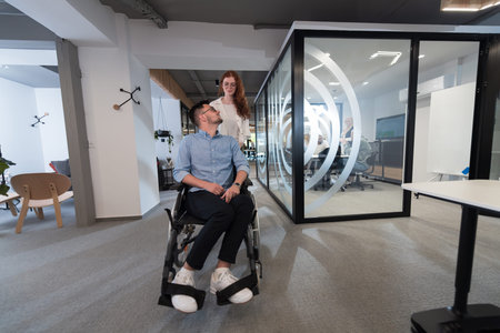 Young business colleagues, collaborative business colleagues, including a person in a wheelchair, walk past a modern glass office corridor, illustrating diversity, teamwork and empowerment in the workplace.の写真素材