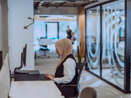 In a modern office, a young Muslim entrepreneur wearing a hijab sits confidently and diligently works on her computer, embodying determination, creativity, and empowerment in the business worldの写真素材