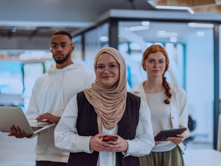 A diverse group of young businessmen, including a Muslim woman wearing a hijab, an orange-haired woman, and an African American, stand together in a modern office, showcasing a vibrant and inclusive workspaceの写真素材