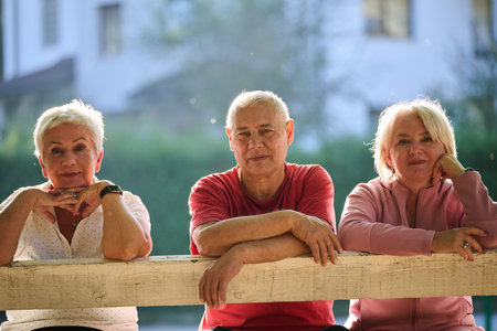 A group of elderly individuals, including a senior man and two older women, sits in a park on a sunny autumn day, embodying the concept of healthy aging through companionship, relaxation, and outdoor vitalityの写真素材