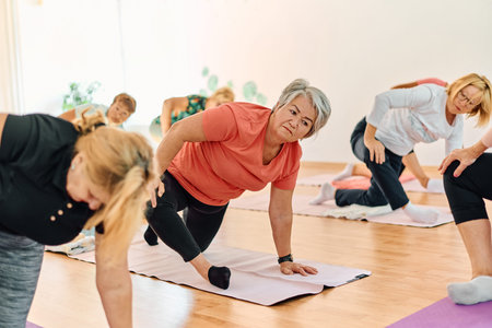 A group of senior women engage in various yoga exercises, including neck, back, and leg stretches,...