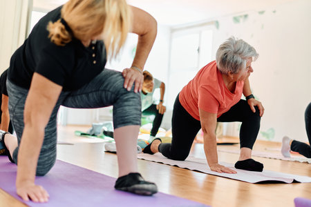 A group of senior women engage in various yoga exercises, including neck, back, and leg stretches, under the guidance of a trainer in a sunlit space, promoting well-being and harmonyの写真素材