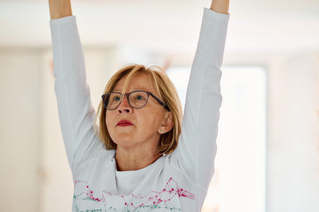 In a sunlit space, a senior woman gracefully practices rejuvenating yoga, focusing on neck, back,...