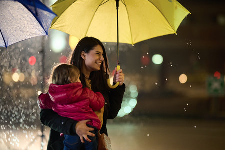 A mother tenderly carries her little daughter while shielding her with an umbrella on a rainy night, embodying the protective love and warmth of maternal care amidst the urban cityscapeの写真素材