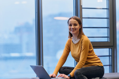 A businesswoman utilizes her laptop while seated by the window of a large corporate building,...