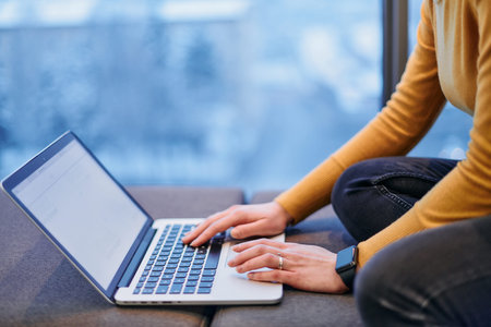 A businesswoman utilizes her laptop while seated by the window of a large corporate building,...