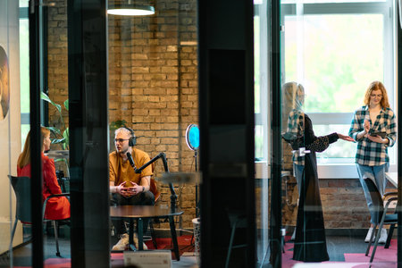 A gathering of young business professionals, some seated in a glass-walled office, engage in a lively conversation and record an online podcast, embodying modern collaboration and dynamic interactionの写真素材