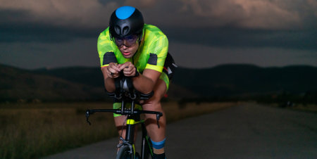 A triathlete rides his bike in the darkness of night, pushing himself to prepare for a marathon. The contrast between the darkness and the light of his bike creates a sense of drama and highlights the athletes determination and perseverance.の写真素材
