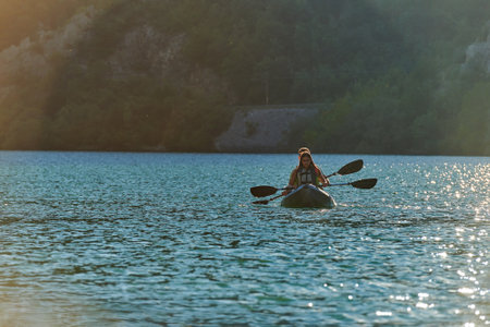 A young couple enjoying an idyllic kayak ride in the middle of a beautiful river surrounded by forest greenery in sunset timeの写真素材