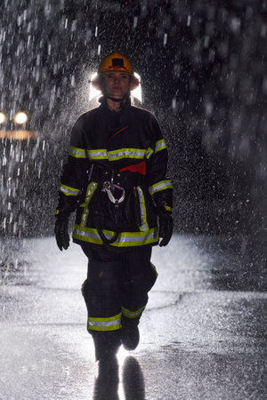A determined female firefighter in a professional uniform striding through the dangerous, rainy night on a daring rescue mission, showcasing her unwavering bravery and commitment to saving lives.の写真素材