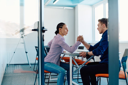 In a modern startup office, a businessman and a businesswoman business colleagues engage in a symbolic arm-wrestling match, reflecting teamwork, competition, and innovation in their dynamic workplace.の写真素材