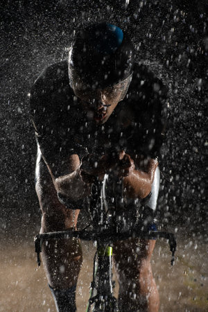 A triathlete braving the rain as he cycles through the night, preparing himself for the upcoming marathon. The blurred raindrops in the foreground and the dark, moody atmosphere in the background add to the sense of determination and grit shown by the athlete.の写真素材