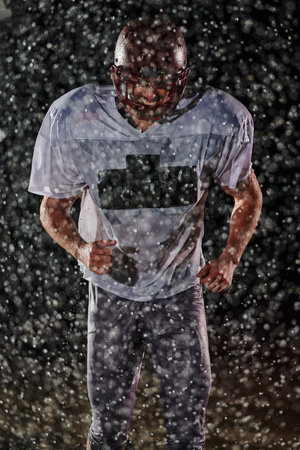 American Football Field: Lonely Athlete Warrior Standing on a Field Holds his Helmet and Ready to Play. Player Preparing to Run, Attack and Score Touchdown. Rainy Night with Dramatic Fog, Blue Lightの写真素材