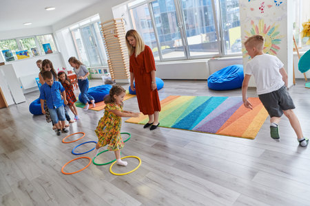 Small nursery school children with female teacher on floor indoors in classroom, doing exercise. Jumping over hula hoop circles track on the floor.の写真素材