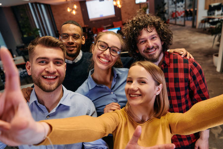 A diverse group of business professionals take a break from their tasks in a modern startup office to capture a creative selfie, showcasing teamwork and a vibrant workplace cultureの写真素材