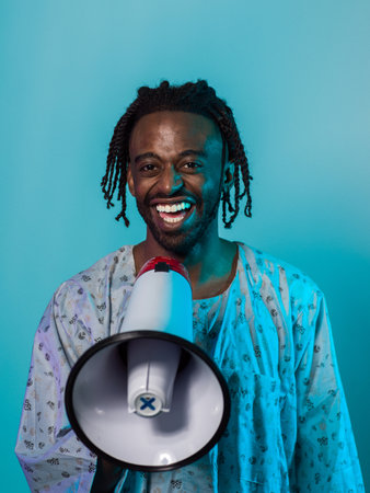 African American man dons traditional attire, passionately utilizing a megaphone against a striking blue background, symbolizing his vocal and cultural empowerment in the pursuit of social justice and equalityの写真素材