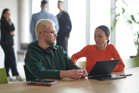 A blond businessman and his colleague sit in an office discussing work while using a laptop to...