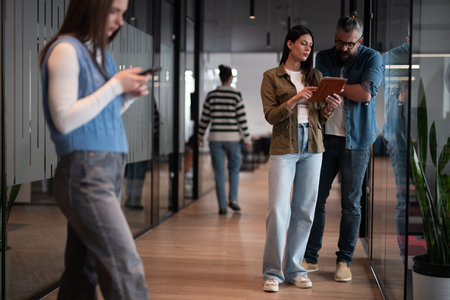 A senior mentor and a leader discuss a project while working on a tablet in a modern coworking space, collaborating confidently while busy colleagues work in the backgroundの写真素材