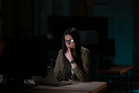 Female businesswoman working late in the office, using a laptop while feeling tired and burnt out. Reading, copywriting, editing, and struggling with fatigue and brain fog on a tight deadlineの写真素材