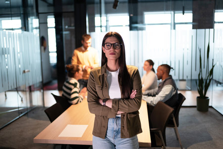 Businesswoman standing confidently in a modern office meeting room. Diverse team on a business meeting in a startup coworking open space office environment collaborating in the background. Corporate teamwork, leadership, and innovationの写真素材