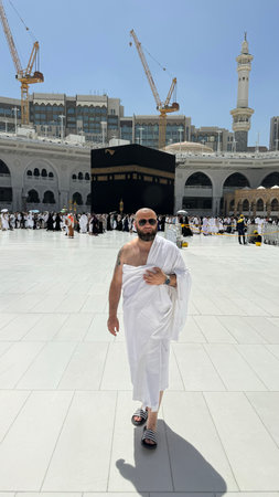 A bald man poses for a photo in front of the Kaaba at noon after performing Umrah at Masjid al-Haram, Mecca. A sacred Islamic pilgrimage in the heart of the holy mosque under the bright sunlight.の写真素材