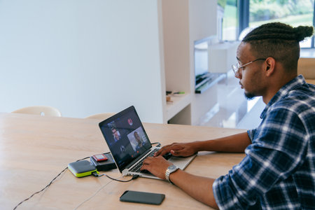 African American influencer in glasses sitting at a table in a modern living room, using a laptop for business video chat, conversation with friends and entertainment.の写真素材