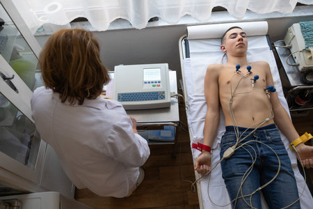 A female doctor performs an ECG on a young man in a hospital room, carefully monitoring his heart health during a routine checkup.の写真素材