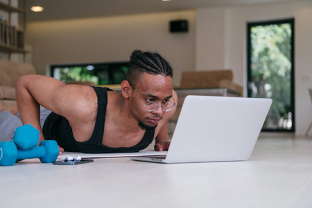Training At Home. Sporty african american man doing the training while watching an online tutorial on a laptop, exercising in the living room, free space.の写真素材