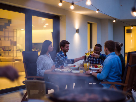 A group of young diverse people having dinner on the terrace of a modern house in the evening. Fun for friends and family. Celebration of holidays, weddings with barbecueの写真素材