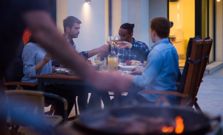A group of young diverse people having dinner on the terrace of a modern house in the evening. Fun for friends and family. Celebration of holidays, weddings with barbecueの写真素材