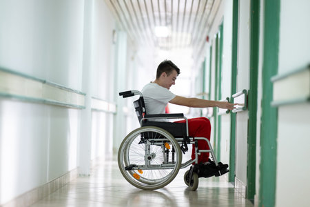 Young teenage boy smiling while sitting in a wheelchair in a hospital hallway. Rehabilitation and...