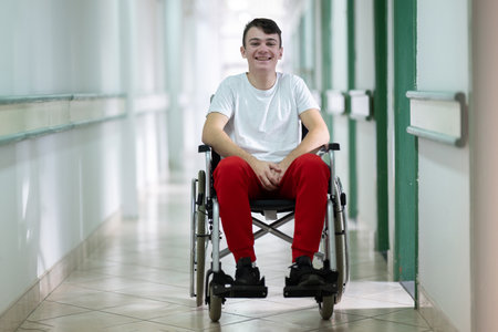 Young teenage boy smiling while sitting in a wheelchair in a hospital hallway. Rehabilitation and positivity concept.の写真素材