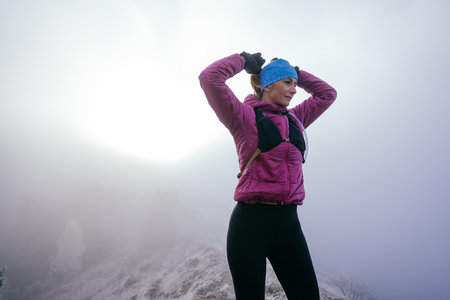 Active female hiker in pink jacket stretching arms on a foggy mountain peak during morning light.の写真素材