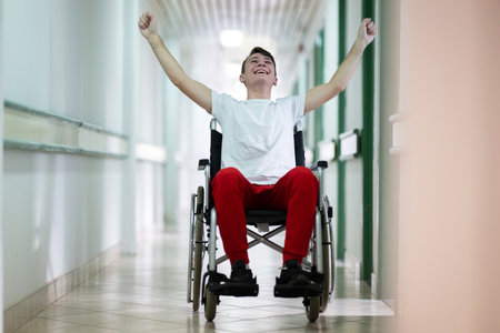 Joyful Young Man in Wheelchair.Teenage boy celebrating in wheelchair with raised arms in hospital hallway. Positivity and recovery.の写真素材