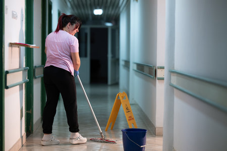 Cleaning Staff in Hospital Hallway.Female janitor standing in a hospital corridor with mop and bucket, symbolizing cleanliness and hygiene.の写真素材