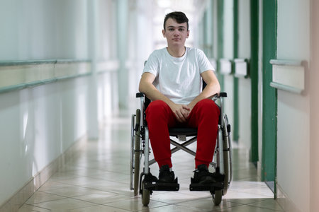 Young teenage boy smiling while sitting in a wheelchair in a hospital hallway. Rehabilitation and positivity concept.の写真素材