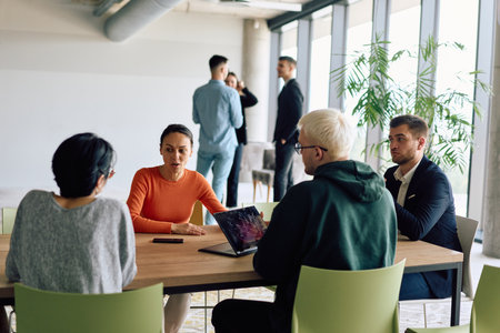 A diverse team of business professionals engaged in a discussion around a conference table in an office, while their colleagues collaborate in the backgroundの写真素材