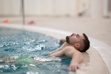 Man floating peacefully in warm spa water with eyes closed and arms outstretched on the edge of the...